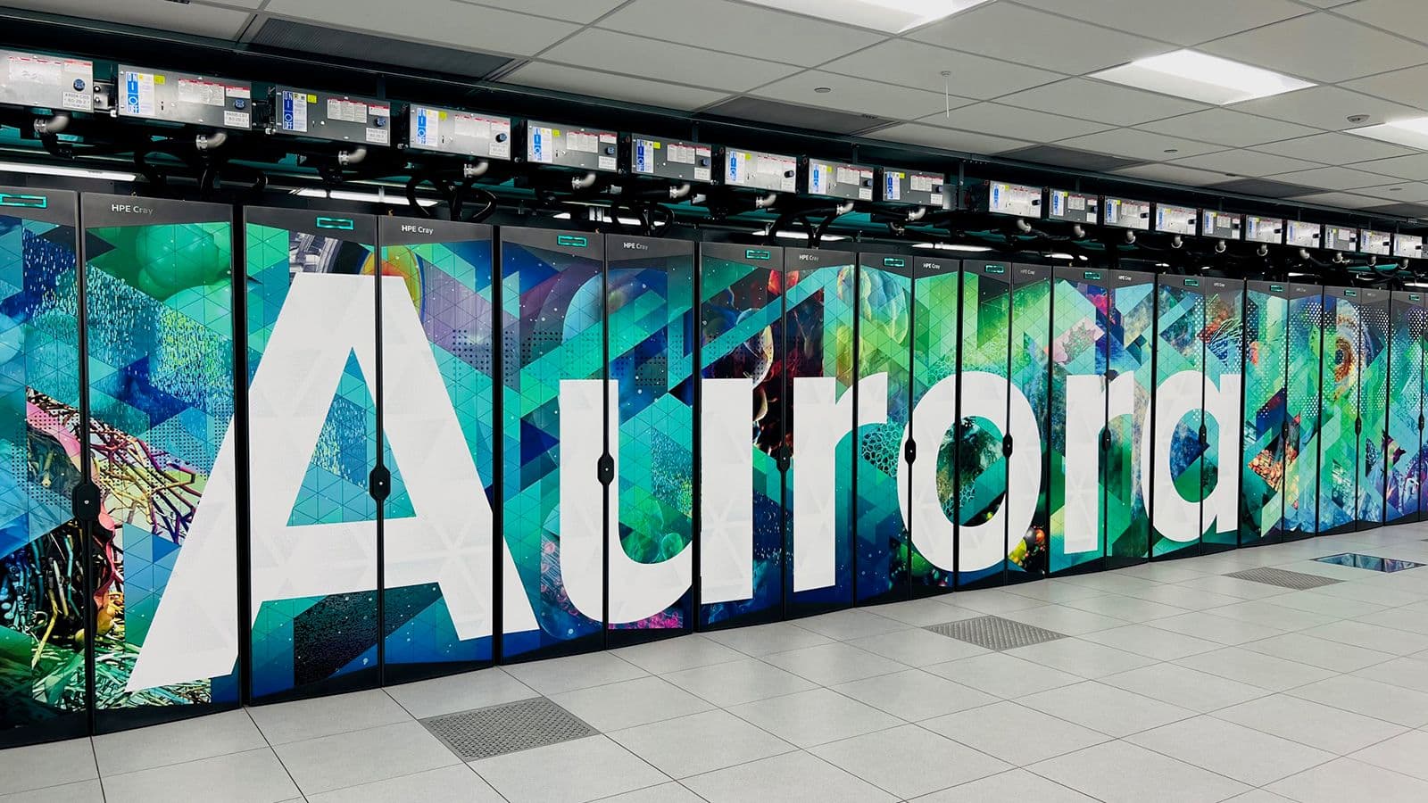 A row of HPE Cray cabinets at Argonne's ALCF spelling "Aurora" in large white letters across teal, green, and magenta panels.