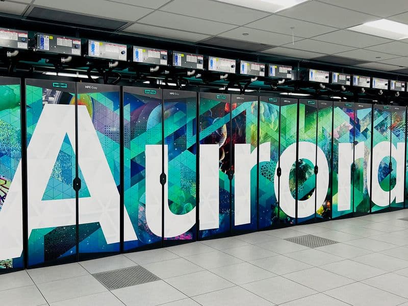A row of HPE Cray cabinets at Argonne's ALCF spelling "Aurora" in large white letters across teal, green, and magenta panels.